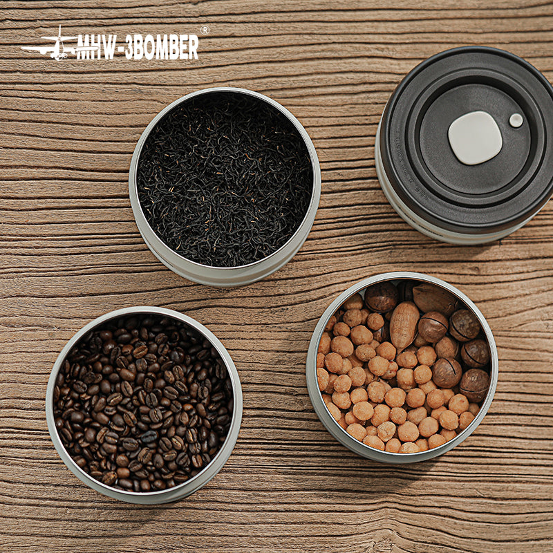 MHW-3BOMBER vacuum canisters with tea, coffee beans, nuts. Round, silver containers on wood surface.