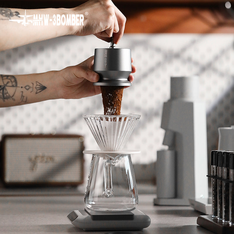 A tattooed individual grinds coffee into a glass pour-over using the MHW-3BOMBER Espresso Coffee Powder Dosing Funnel. Ideal for enthusiasts and baristas, the setup includes a scale beneath it. Blurred kitchen items and decor are visible in the background.