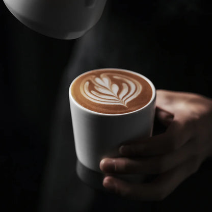 MHW-3BOMBER CERA white ceramic cup with latte art being held. U-shaped for aroma. Heat-resistant. 