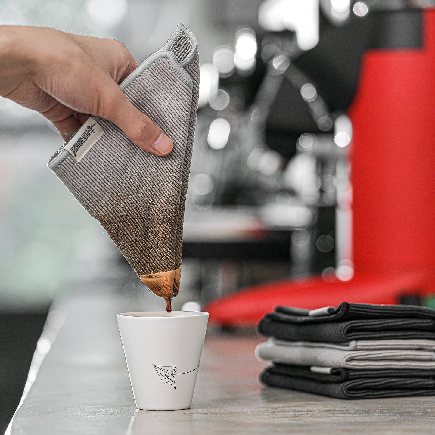 MHW-3BOMBER Barista Towel Set. Grey cloth coffee filter in use over a cup. Stack of towels beside.