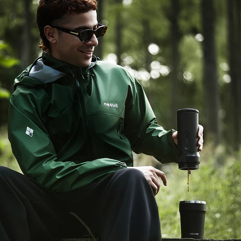 Person in green jacket sitting outdoors, pouring coffee from a black travel mug.