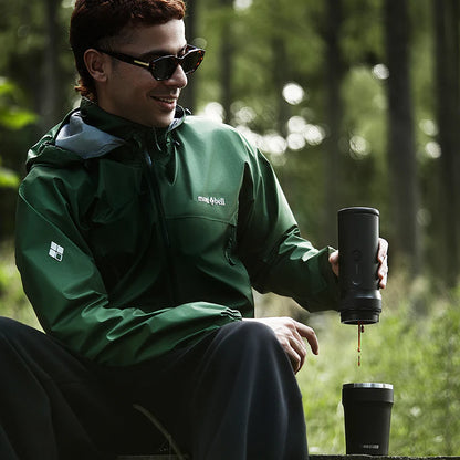 Person in green jacket sitting outdoors, pouring coffee from a black travel mug.