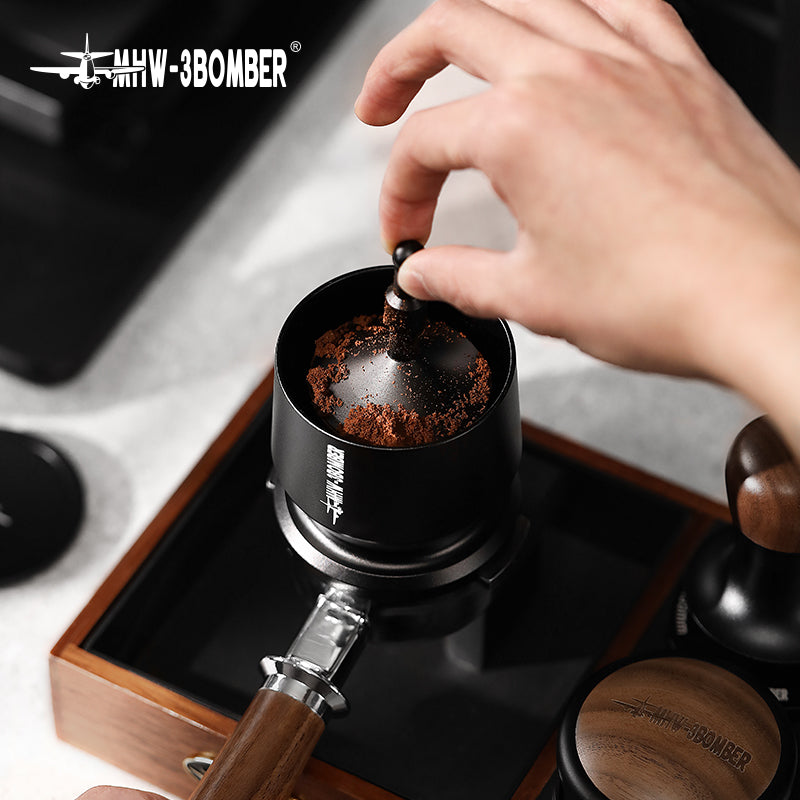 Hands using the MHW-3BOMBER Espresso Coffee Powder Dosing Funnel to evenly distribute ground coffee in a portafilter, ensuring precision. Nearby are a wooden handle tamper and box, with MHW-3BOMBER branding at the top left—ideal for baristas or home enthusiasts.
