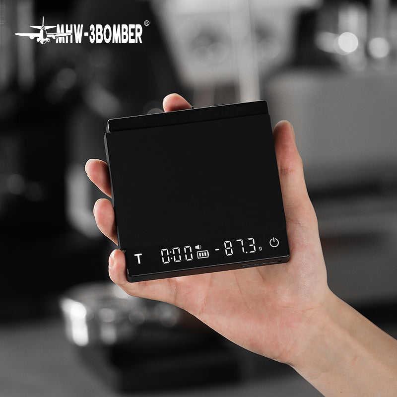 A hand holds the MHW-3BOMBER Cube Coffee Scale 2.0 Mini, featuring a black surface and clear display. This USB rechargeable digital scale is set against a blurred interior, suggesting a kitchen or coffee setting.