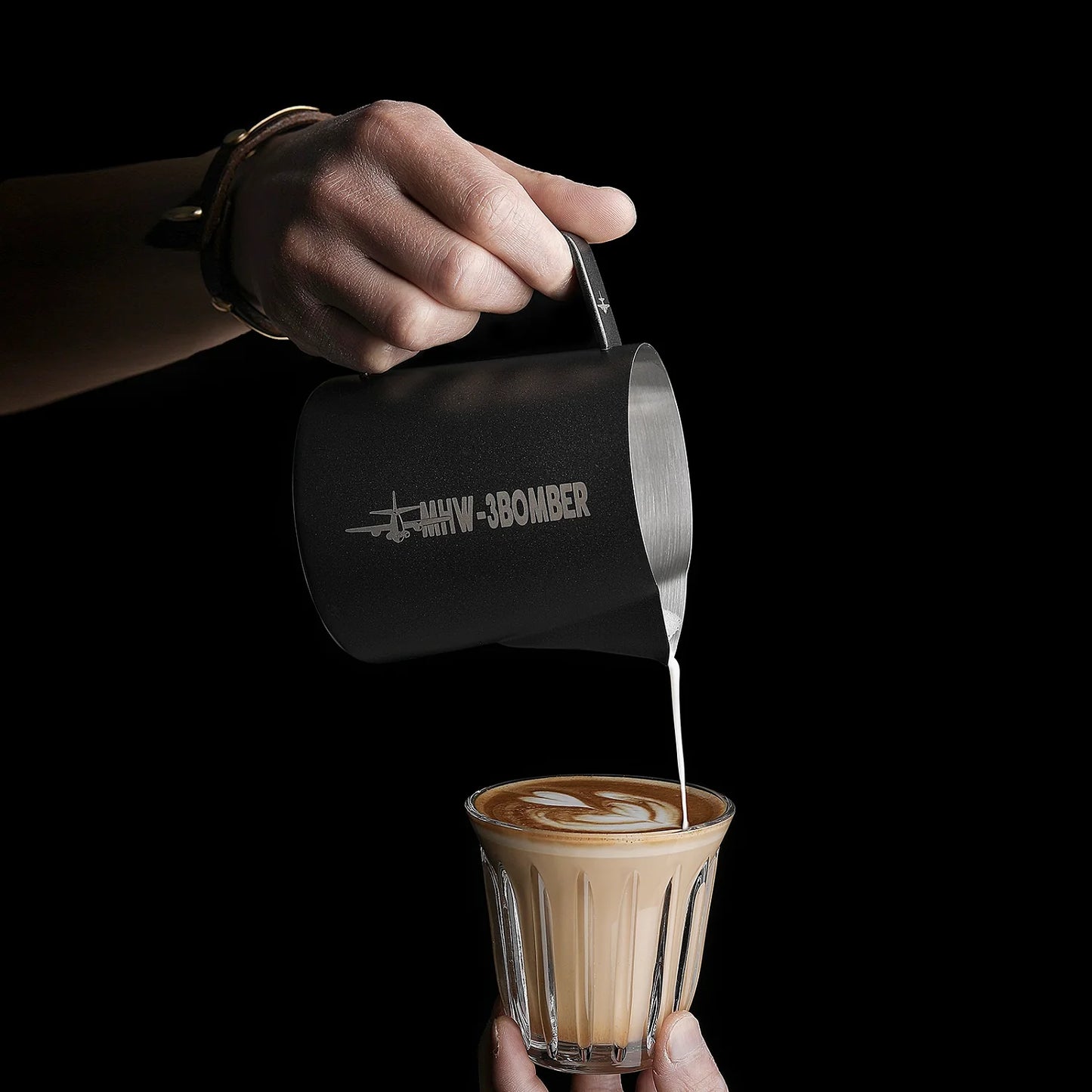 A hand holds a matte black MHW-3BOMBER Latte Art Pitcher 5.0 with a crocodile spout, skillfully pouring steamed milk into a glass of coffee below to create latte art.