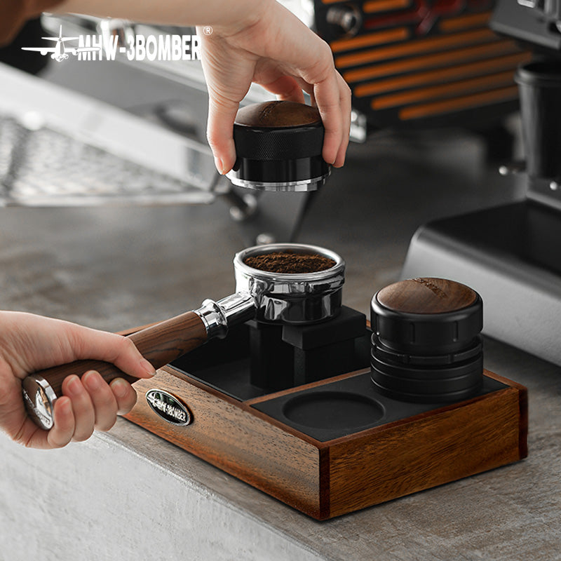 MHW-3BOMBER espresso tamping station with portafilter, tamper, and wooden base. Espresso accessories on a kitchen counter.