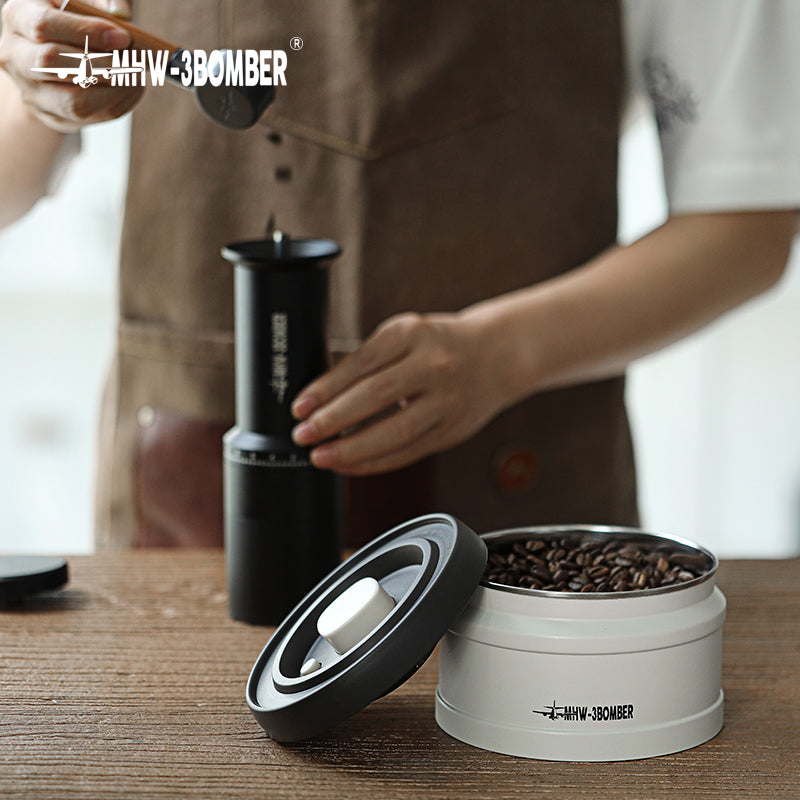 MHW-3BOMBER vacuum canister (white) and coffee grinder (black) with beans, on wood table. 