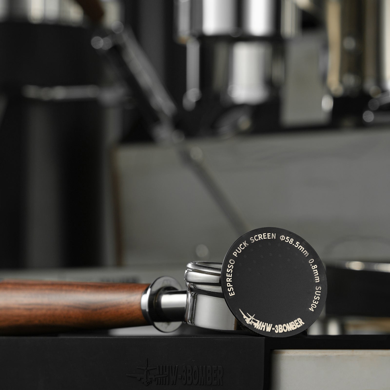 A wooden handle portafilter rests on a table with a close-up of the double-layer MHW-3BOMBER Espresso Puck Screen in titanium plated black, featuring its dimensions and brand name, set against a blurred coffee machine backdrop.