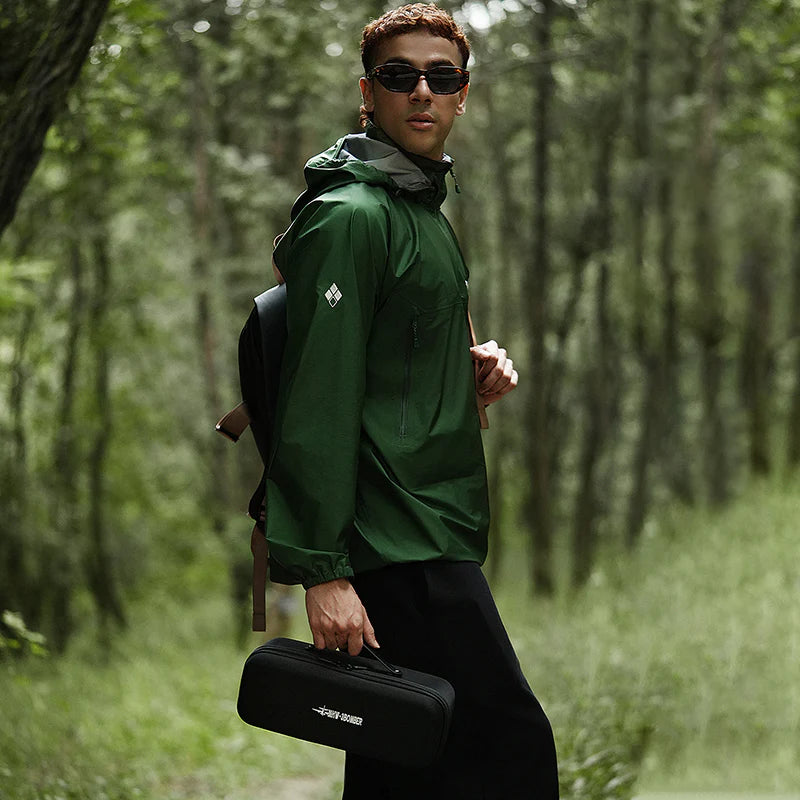 Man in green jacket and sunglasses holding a black case in a forest