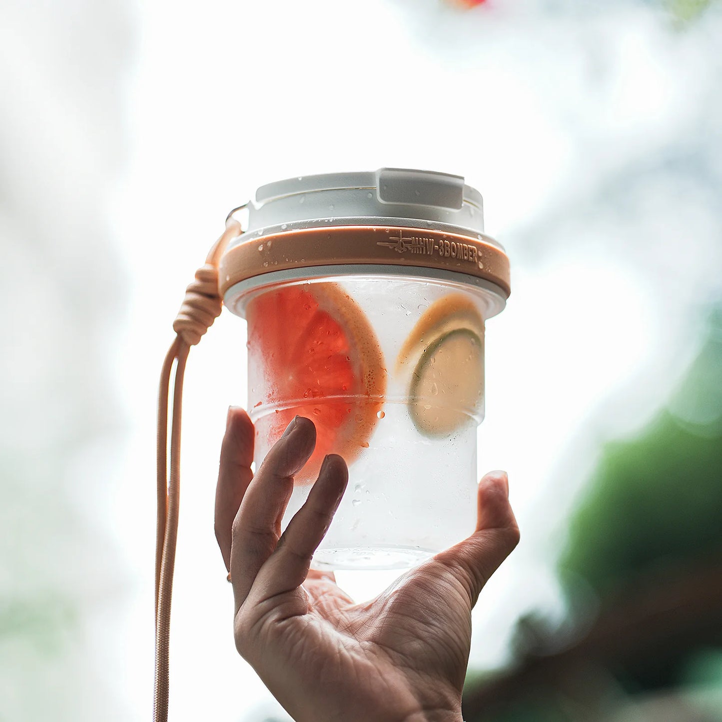 MHW-3BOMBER portable cup with fruit. Clear body, tan accents, and strap. Leak-proof design. Held in a hand.