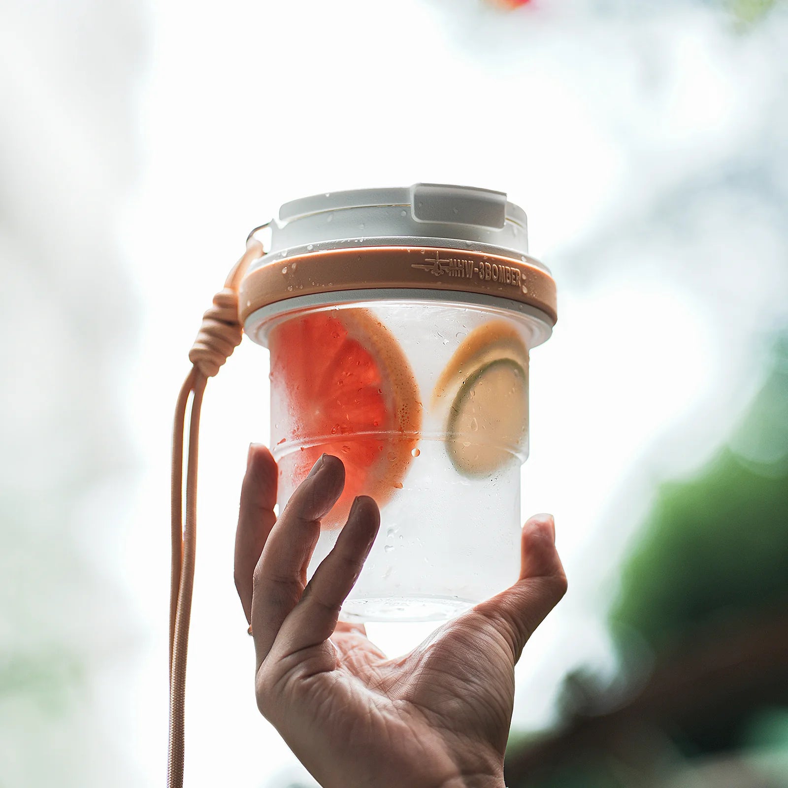 MHW-3BOMBER portable cup with fruit. Clear body, tan accents, and strap. Leak-proof design. Held in a hand.