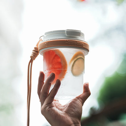 MHW-3BOMBER portable cup with fruit. Clear body, tan accents, and strap. Leak-proof design. Held in a hand.