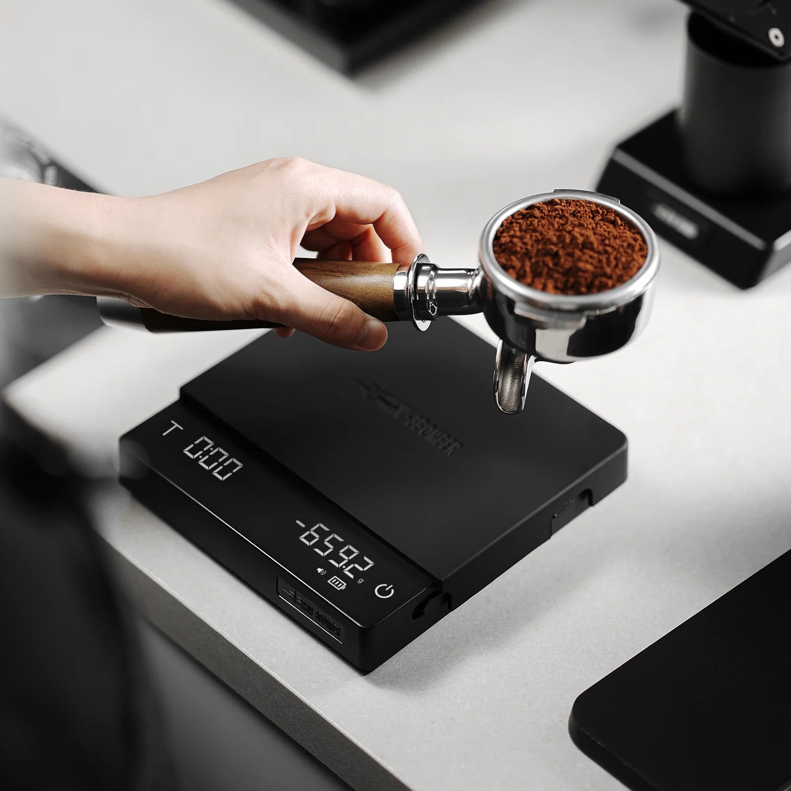 MHW-3BOMBER black cube coffee scale with portafilter and grounds on top. Digital display visible.