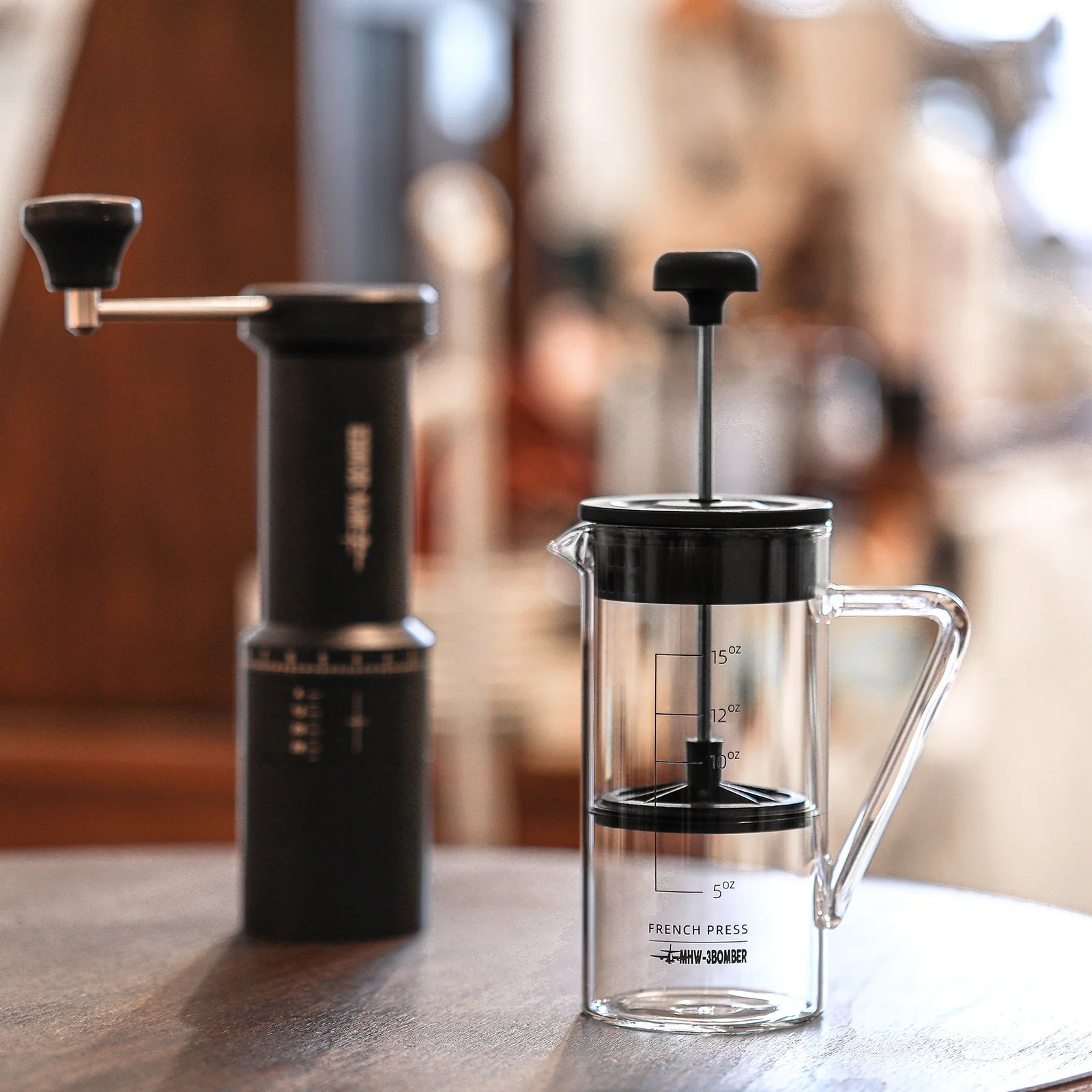 The MHW-3BOMBER Gimme French Press, made with heat-resistant glass and a black handle, sits on a wooden table by a black manual coffee grinder. With measurement markings, it adds to the cozy atmosphere of the blurred background.