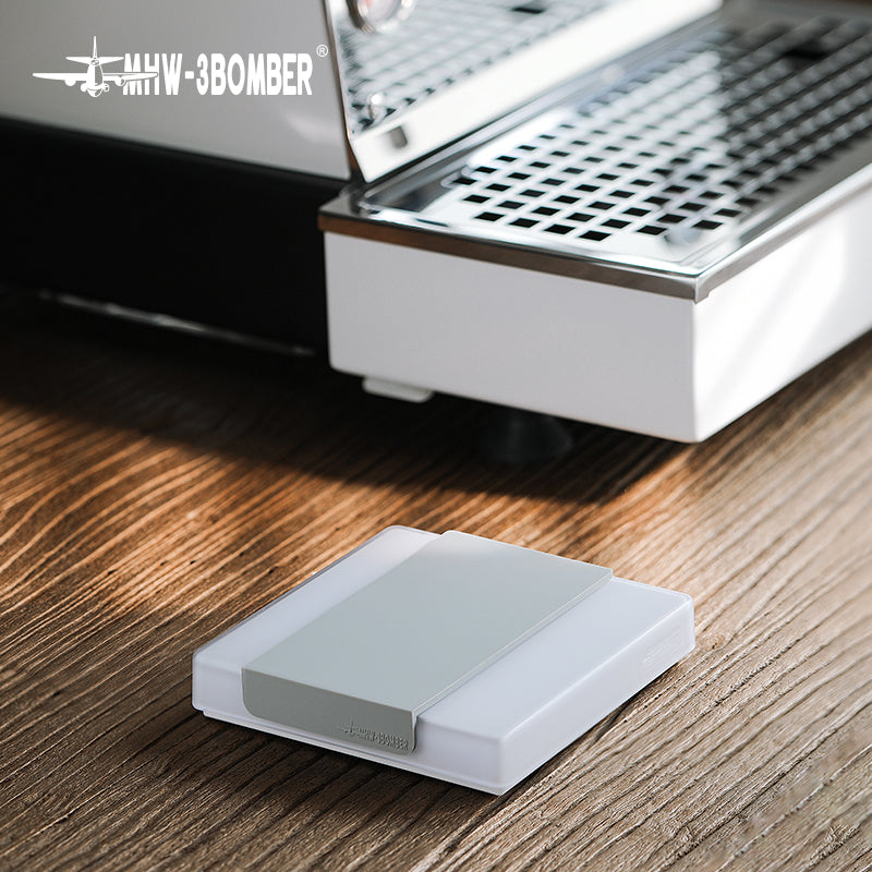 On a wooden surface beside a modern metal appliance with a grid design, theres a white rectangular MHW-3BOMBER Cube Coffee Scale 2.0 Mini featuring a gray band and the brands logo in the top left corner.
