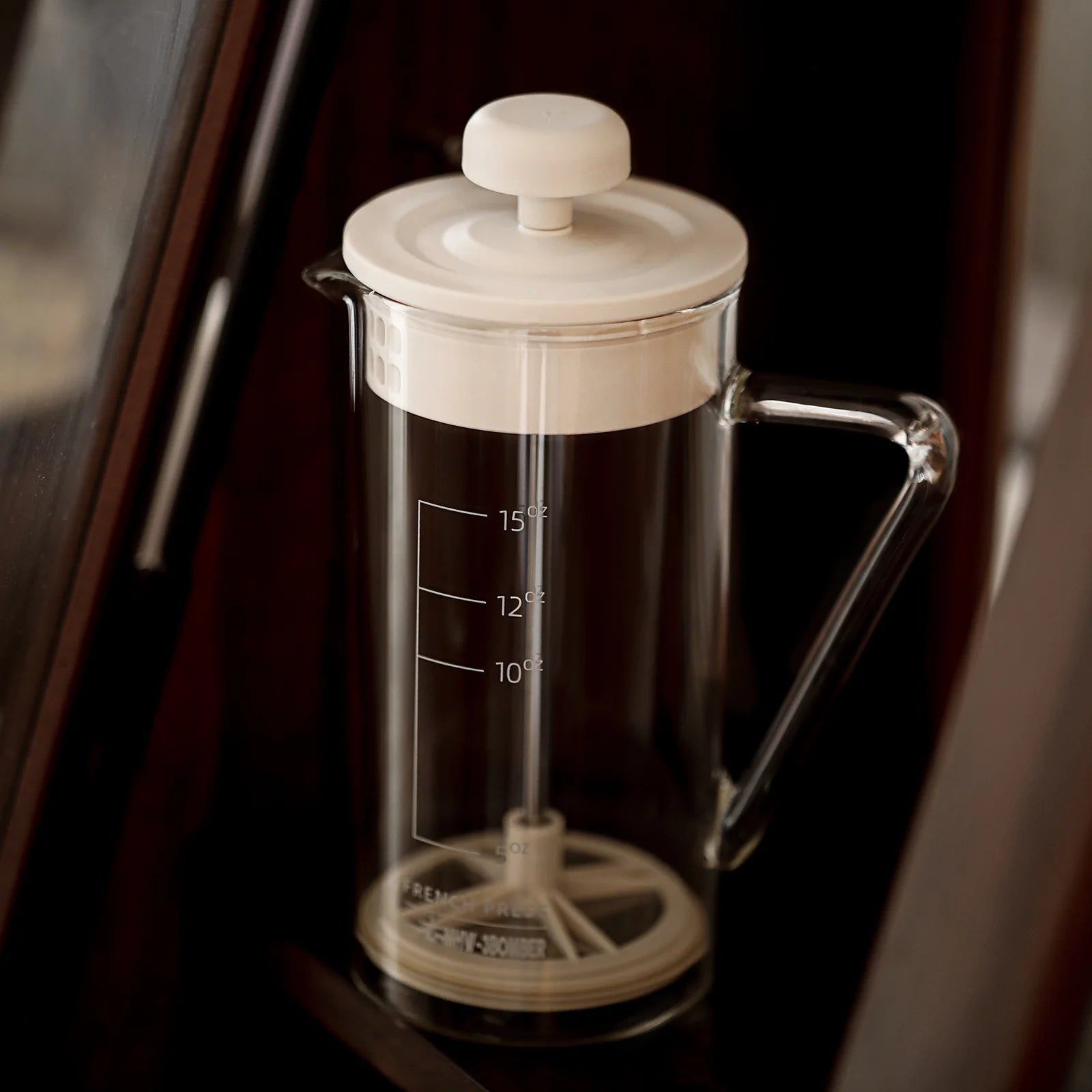 The MHW-3BOMBER Gimme French Press features a heat-resistant, clear glass body with a white lid and plunger. It includes measurement markings, rests on a dark surface, and boasts an elegant design with a stainless steel filter.