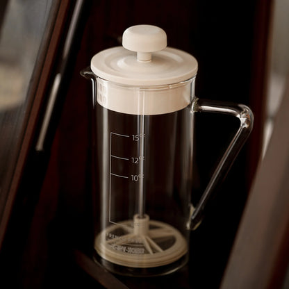 The MHW-3BOMBER Gimme French Press features a heat-resistant, clear glass body with a white lid and plunger. It includes measurement markings, rests on a dark surface, and boasts an elegant design with a stainless steel filter.