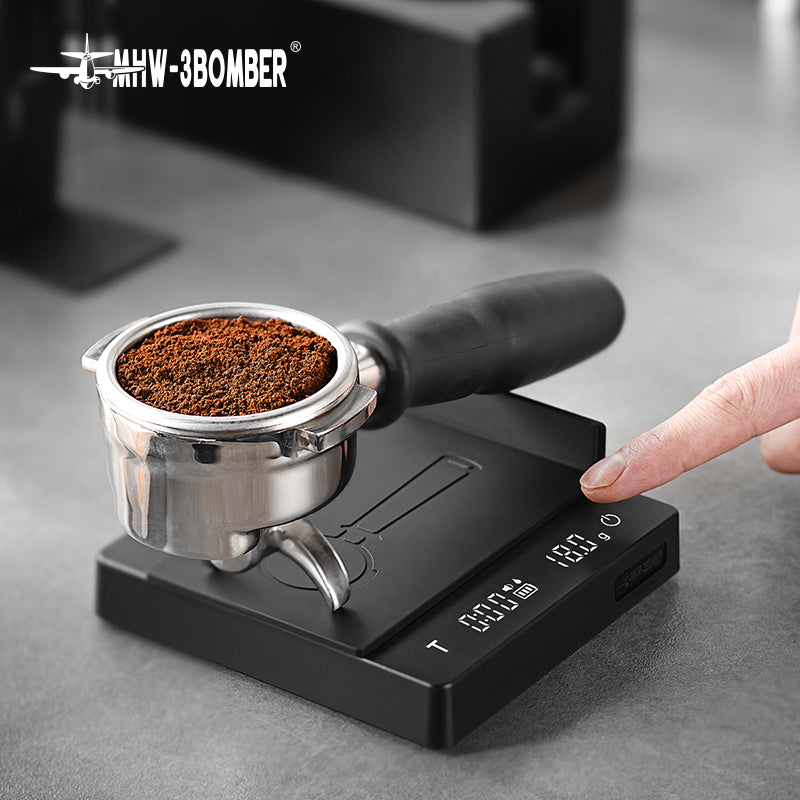 A hand presses a button on the MHW-3BOMBER Cube Coffee Scale 2.0 Mini, with a portafilter of ground coffee on the USB rechargeable scale. The weight and timer display are visible on the gray countertop.