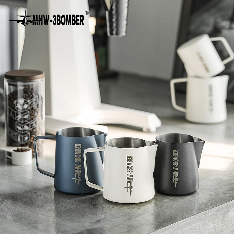 On a minimalist kitchen counter, three MHW-3BOMBER pitchers in blue, white, and black sit elegantly. Crafted from stainless steel and featuring crocodile spouts for precision pouring, they accompany a coffee grinder and stacked mugs, creating a modern scene.