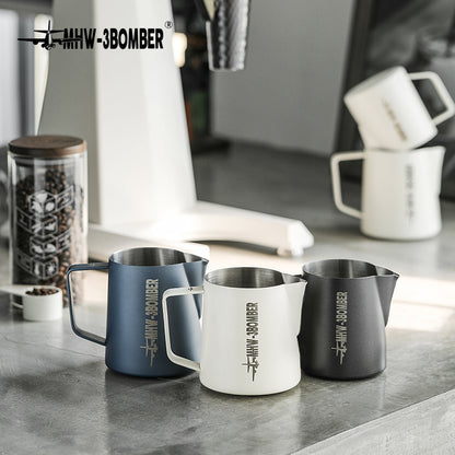 On a minimalist kitchen counter, three MHW-3BOMBER pitchers in blue, white, and black sit elegantly. Crafted from stainless steel and featuring crocodile spouts for precision pouring, they accompany a coffee grinder and stacked mugs, creating a modern scene.