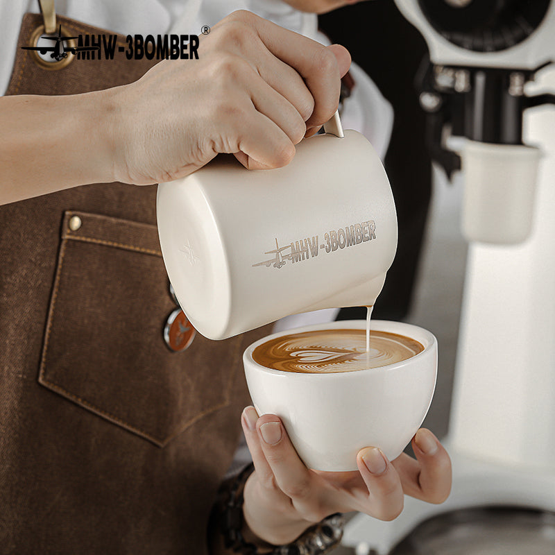 A person pours latte art into a coffee cup using the MHW-3BOMBER Latte Art Pitcher 5.0 White, wearing a brown apron, crafting drinks with precision. In the background are a coffee machine and grinder, setting the perfect scene for this baristas artistry.