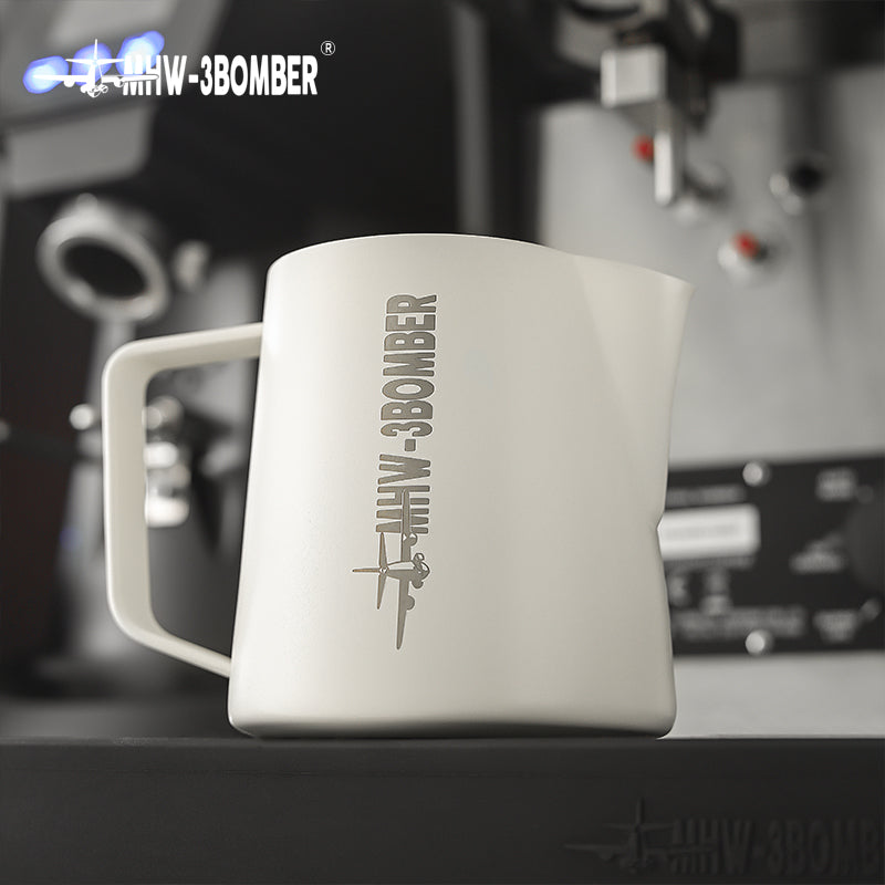 A MHW-3BOMBER Latte Art Pitcher 5.0 White sits on a countertop, while a stainless steel milk frother is visible next to coffee-making machinery in the background.