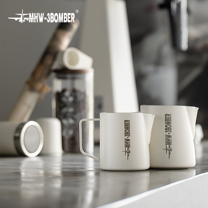 Two white MHW-3BOMBER Latte Art Pitcher 5.0 mugs rest on a reflective surface, with a blurry coffee canister and other kitchen items, like a sleek stainless steel milk frother, enhancing the background ambiance.