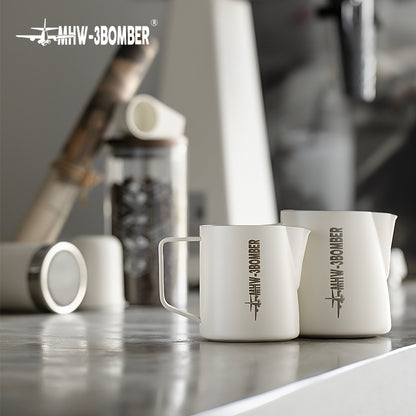Two white MHW-3BOMBER Latte Art Pitcher 5.0 mugs rest on a reflective surface, with a blurry coffee canister and other kitchen items, like a sleek stainless steel milk frother, enhancing the background ambiance.