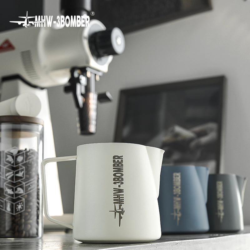 The MHW-3BOMBER Latte Art Pitcher 5.0 in white, featuring a precise crocodile spout, is in focus. In the background are a coffee grinder and two blue pitchers, with a blurred picture hanging on the wall.