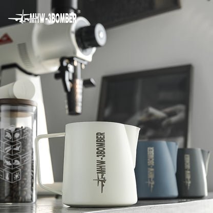 The MHW-3BOMBER Latte Art Pitcher 5.0 in white, featuring a precise crocodile spout, is in focus. In the background are a coffee grinder and two blue pitchers, with a blurred picture hanging on the wall.