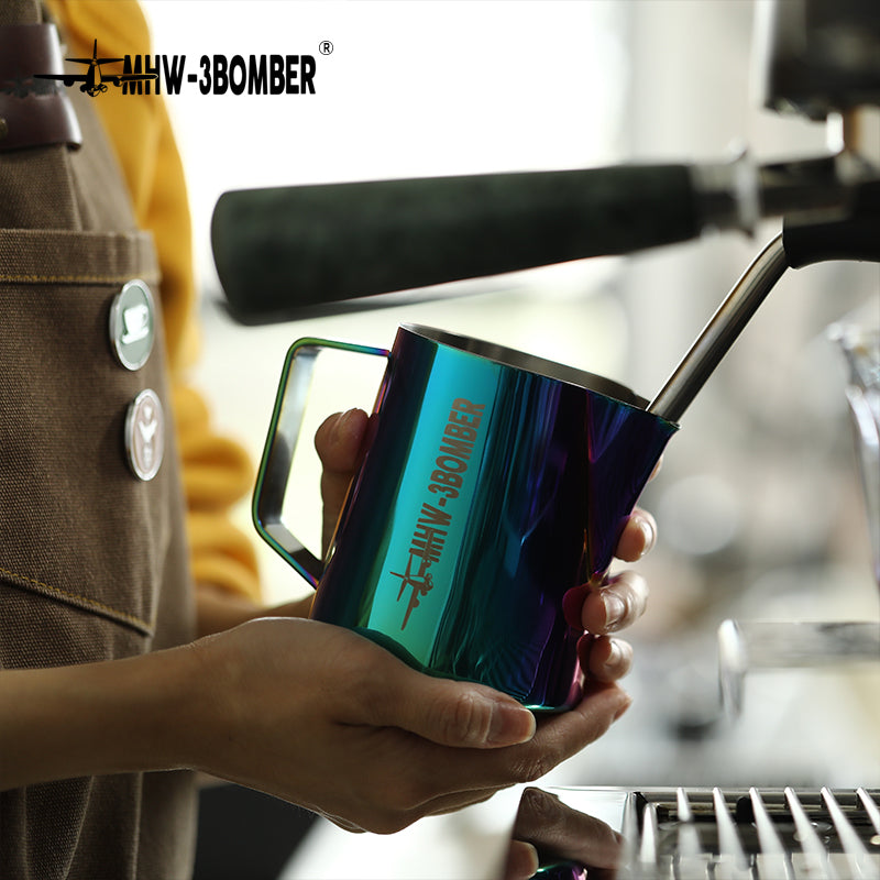 In a coffee shop, a barista in a brown apron holds the colorful MHW-3BOMBER Latte Art Pitcher 5.0 with a crocodile spout beside a steaming wand.