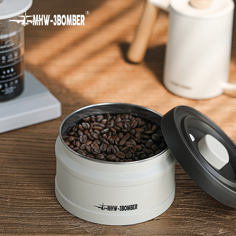 MHW-3BOMBER vacuum coffee canister with beans. White container, open lid showing contents on table.