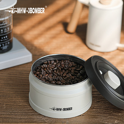 MHW-3BOMBER vacuum coffee canister with beans. White container, open lid showing contents on table.