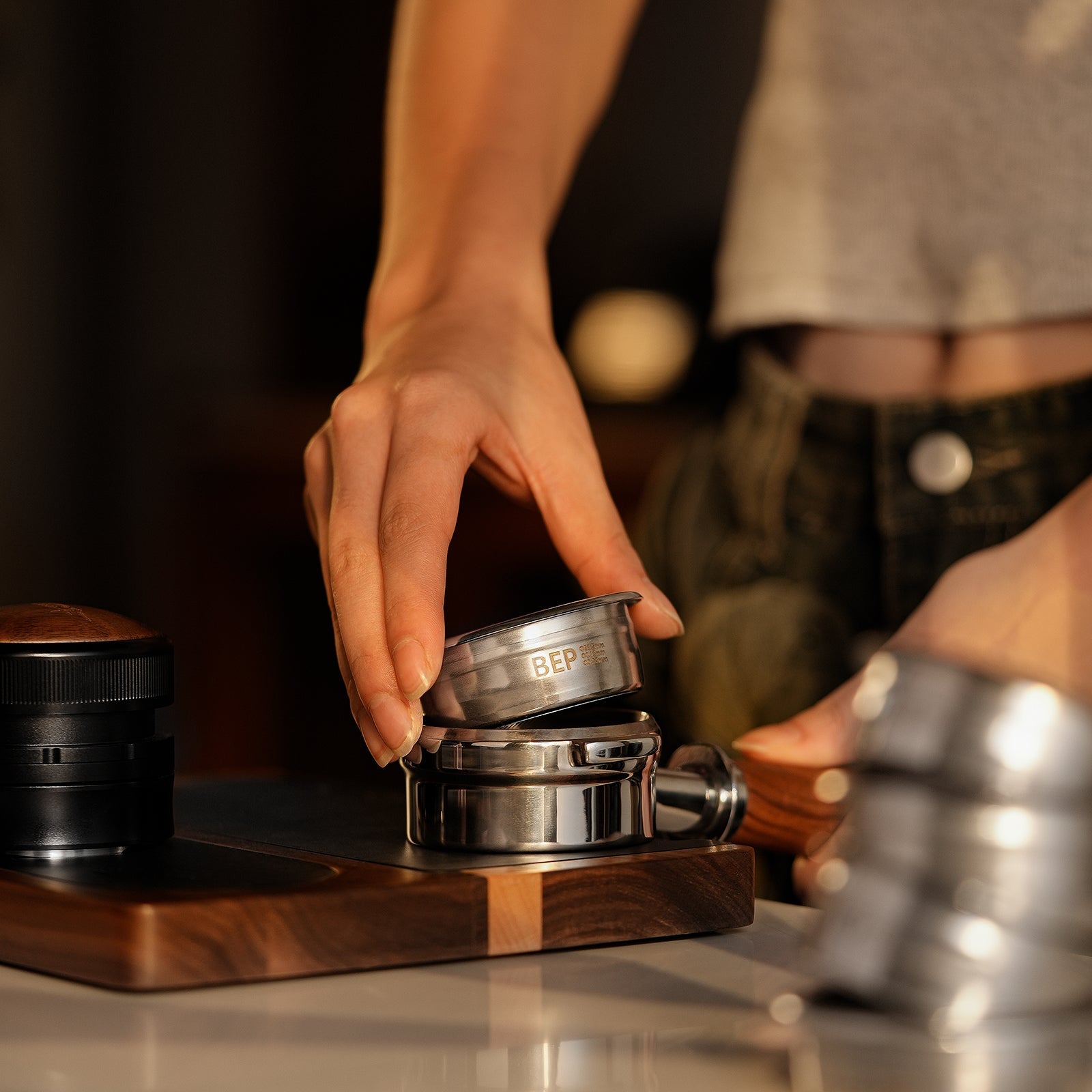 Wearing a light top and jeans, a person uses an MHW-3BOMBER BEP Butterfly Coffee Filter Basket 2.0 (58.5mm) with an espresso machine portafilter in a warmly lit kitchen or coffee bar.