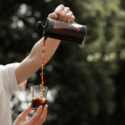 A person pours coffee from the stylish MHW-3BOMBER Gimme French Press with a stainless steel filter into a small heat-resistant glass. The scene, set outdoors amid blurred green foliage, captures the mid-pour moment in natural light for a dynamic effect.