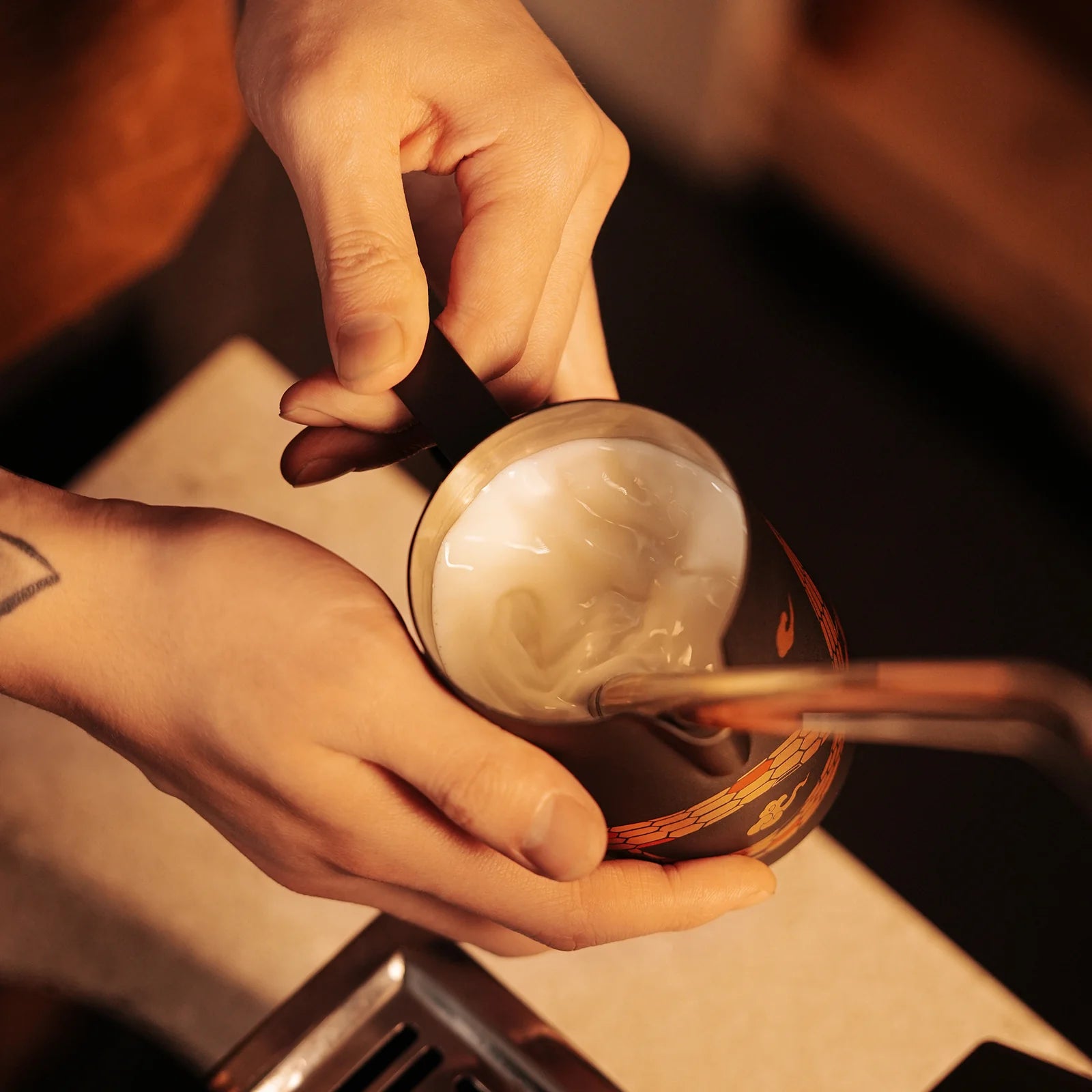 A close-up shows hands using the MHW-3BOMBER Turbo Milk Pitcher-Year of the Snake Limited Edition, stirring frothy milk. A wrist tattoo adds personal style in the warmly lit setting, blending tradition and modern charm.
