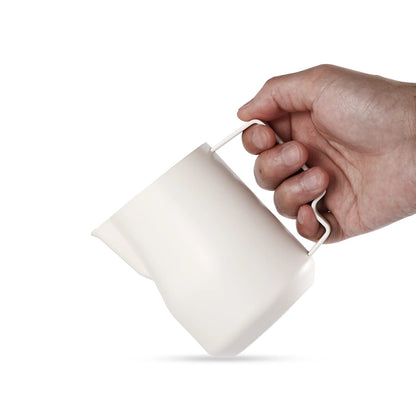 A hand holds the MHW-3BOMBER Turbo Milk Pitcher White, featuring a distinctive shape with a narrow handle and angular spout, ideal for milk frothing. The pitcher is displayed sideways against a plain white background.