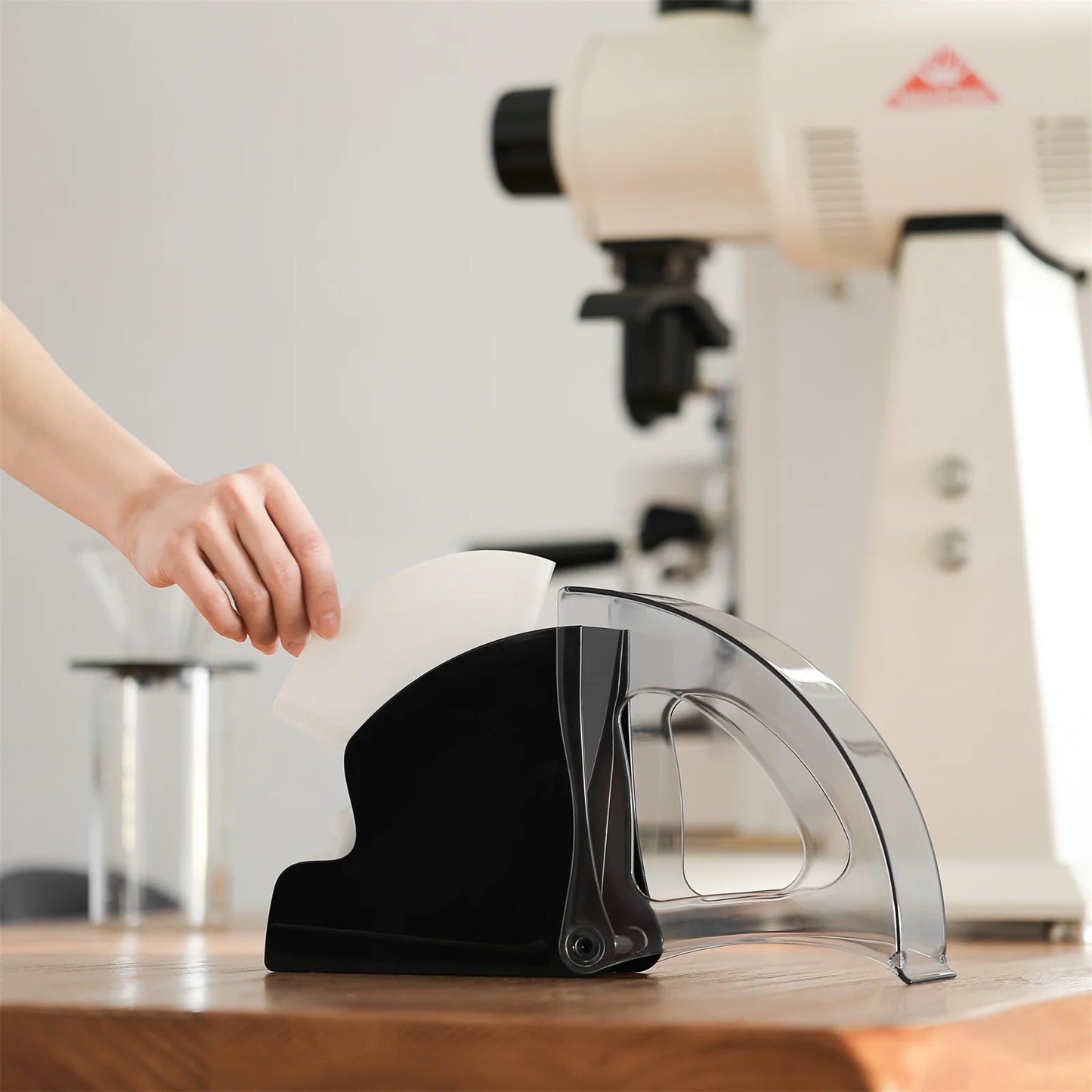 MHW-3BOMBER black coffee filter holder with clear lid and paper filter, on wood counter with grinder in background.