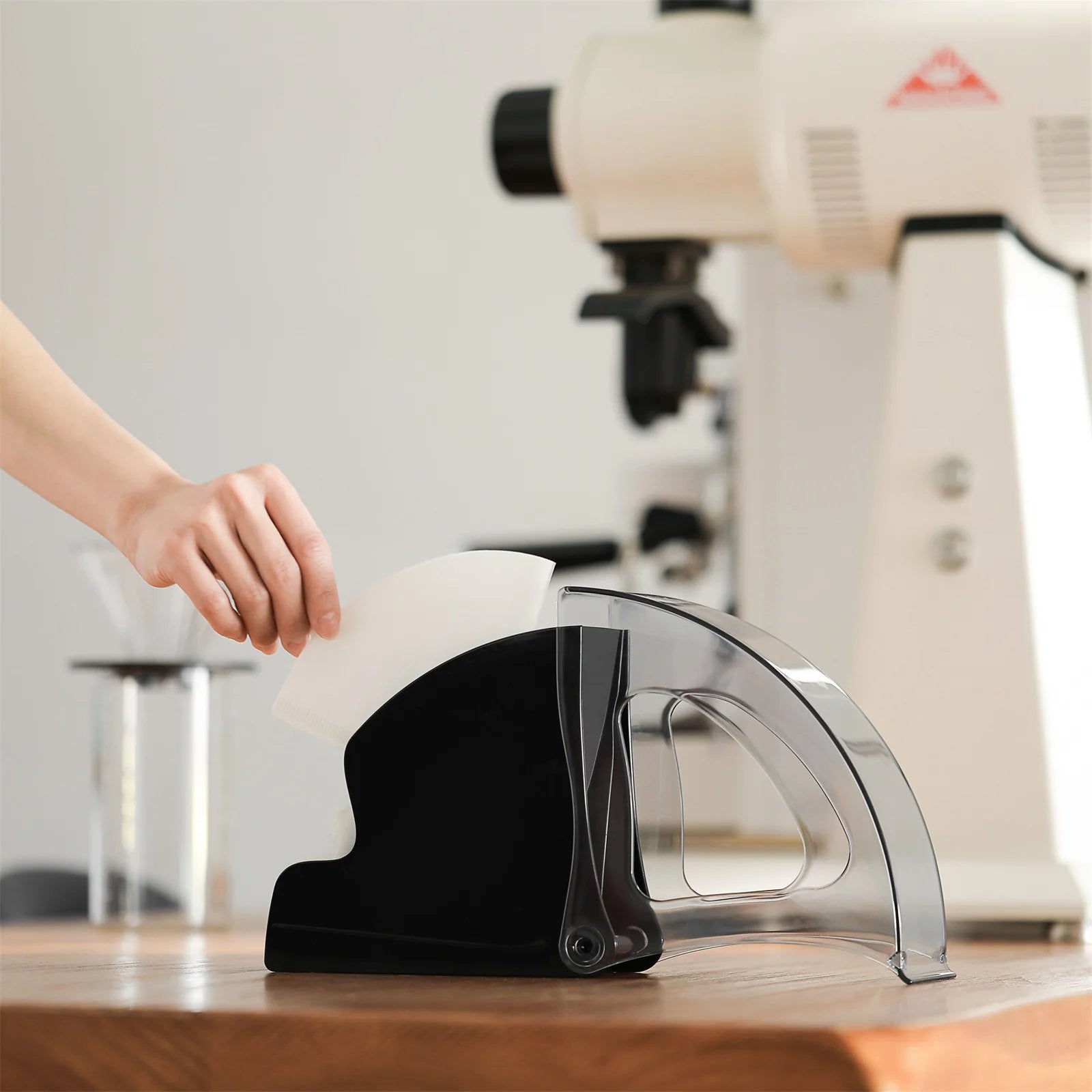 MHW-3BOMBER black coffee filter holder with clear lid and paper filter, on wood counter with grinder in background.