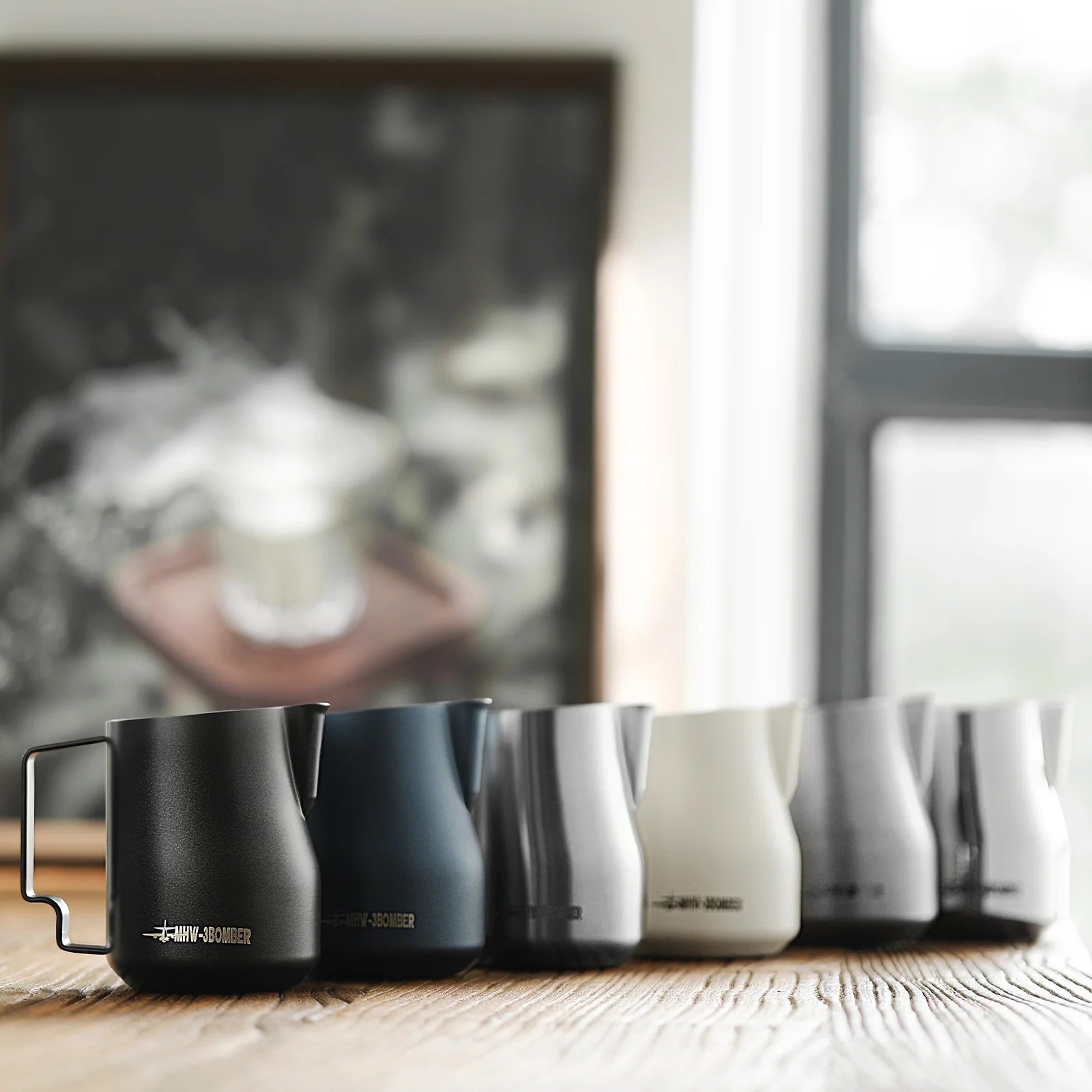 A row of six MHW-3BOMBER Turbo Milk Pitcher Glossy pitchers in black, blue, gray, white, silver, and a light shade rest on a wooden surface. These stainless steel pitchers are ideal for creating latte art. A window and framed picture blur in the background.