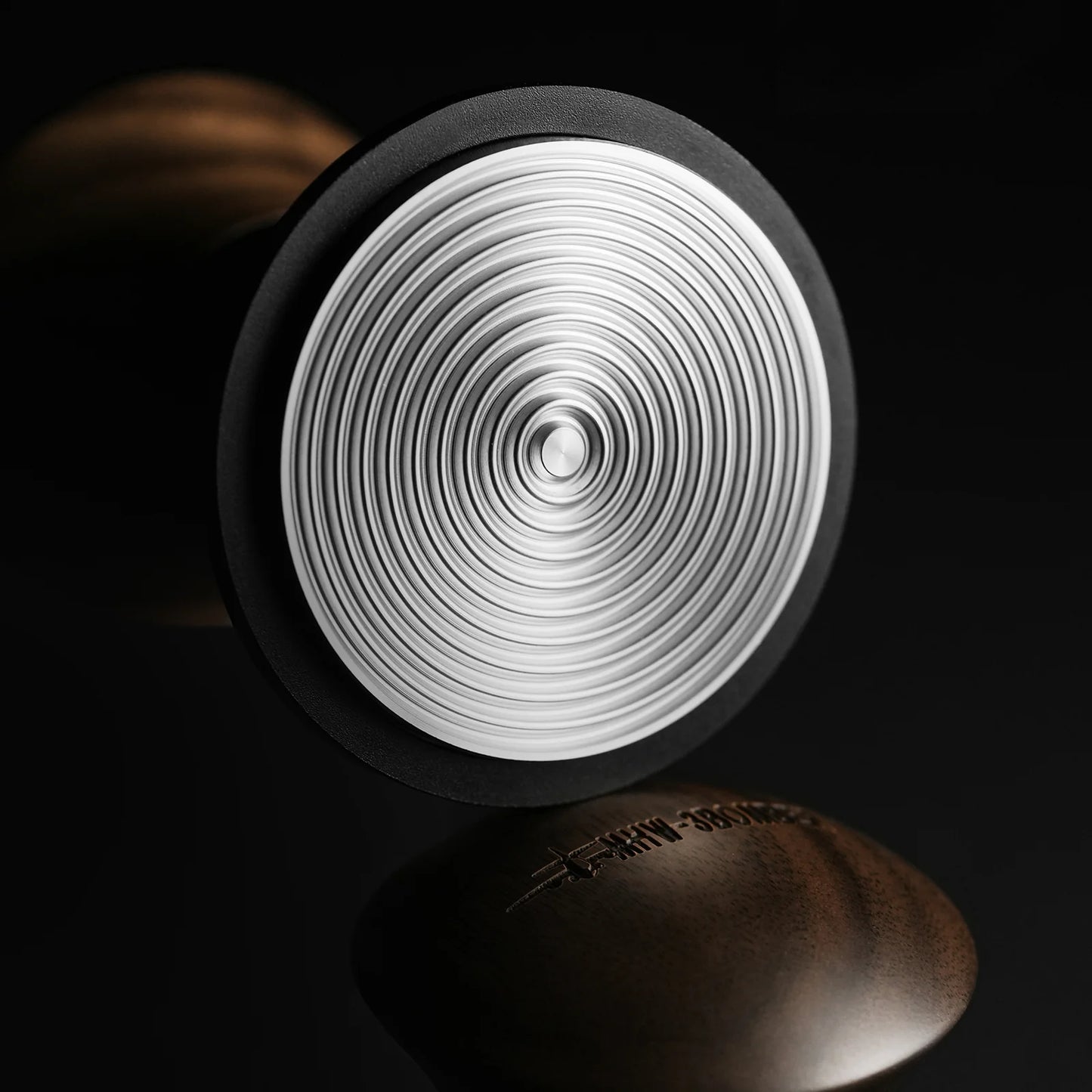 A close-up shows the MHW-3BOMBER Armor Series Coffee Tamper 2.0 with a textured concentric circle pattern on its silver base against a black backdrop, featuring adjustable tamping depth. The blurred logo is visible on the handles side in the foreground.