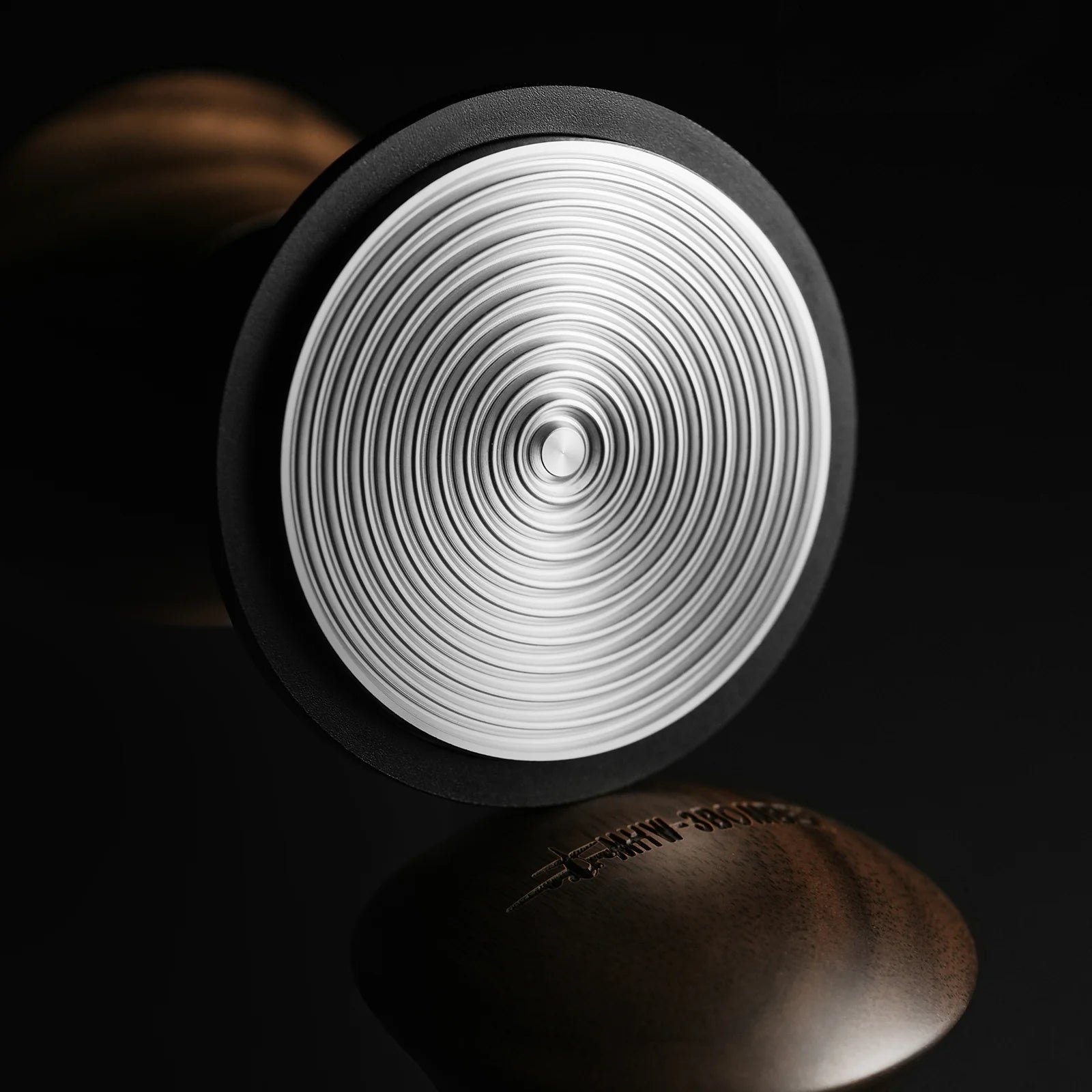 A close-up shows the MHW-3BOMBER Armor Series Coffee Tamper 2.0 with a textured concentric circle pattern on its silver base against a black backdrop, featuring adjustable tamping depth. The blurred logo is visible on the handles side in the foreground.