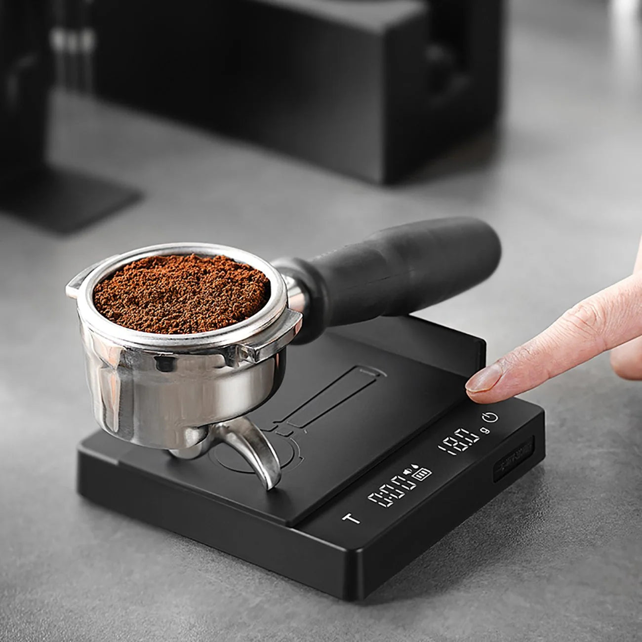 A portafilter with ground coffee sits on the MHW-3BOMBER Espresso Scale Cube Coffee Scale 2.0, ready for a finger to press the button as numbers glow on its digital display. This sleek, USB-rechargeable device rests elegantly on a smooth gray surface.