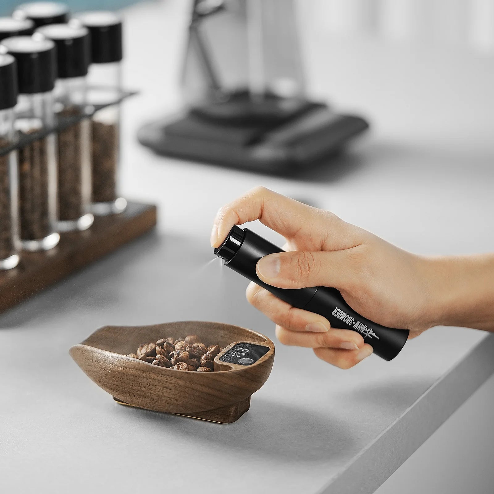 A hand holds the MHW-3BOMBER Ovo Coffee Scale & Spray Bottle, spritzing onto coffee beans in a wooden bowl on a gray counter, enhancing the brewing process with test tubes of spices in the background.