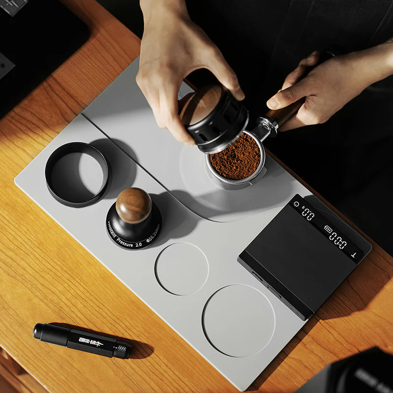 MHW-3BOMBER gray silicone tamping base with portafilter, tamper, WDT tool, dosing ring, and scale. Coffee prep on wood counter.