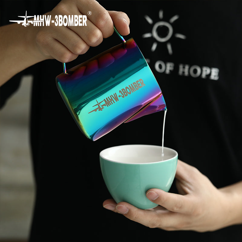 A person pours liquid from a metallic, iridescent MHW-3BOMBER Latte Art Pitcher 5.0 with a crocodile spout into a light blue ceramic cup. The pitcher features an airplane logo and MHW-3BOMBER text. The person wears a casual black t-shirt with a sun design and text.