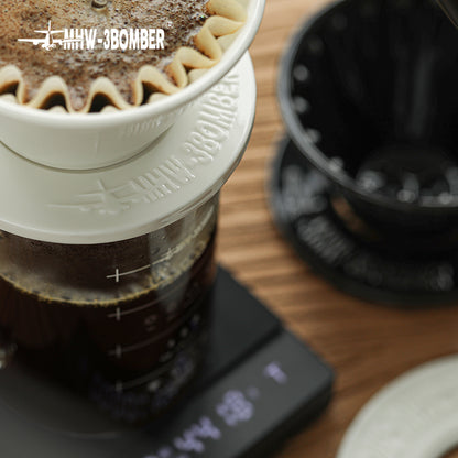 MHW-3BOMBER Meteor coffee dripper on scale. White ceramic pour-over brewing. Black coffee in glass container.