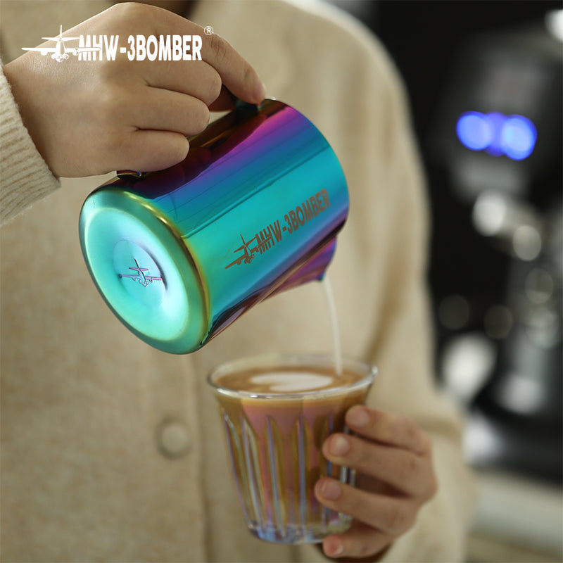 A person in a beige sweater is pouring milk from a colorful MHW-3BOMBER Latte Art Pitcher, featuring a stainless steel body and crocodile spout, into a glass of coffee.
