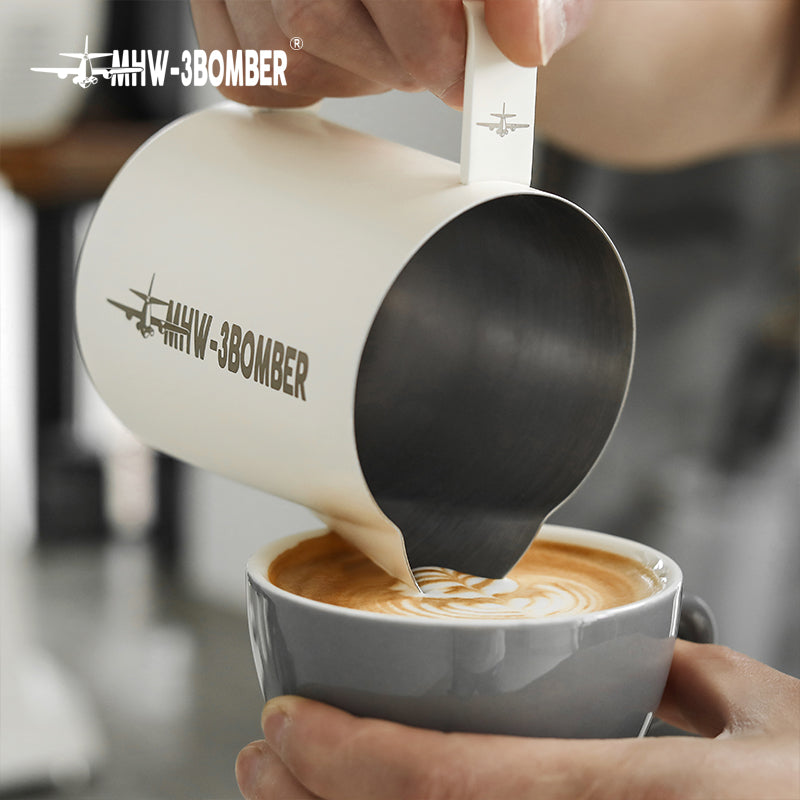 A person pours milk into a coffee cup through a crocodile spout from the MHW-3BOMBER Latte Art Pitcher 5.0 White, adding style with its branded airplane logo. The coffee cup remains steady on the saucer, elegantly capturing the intricate latte art design.
