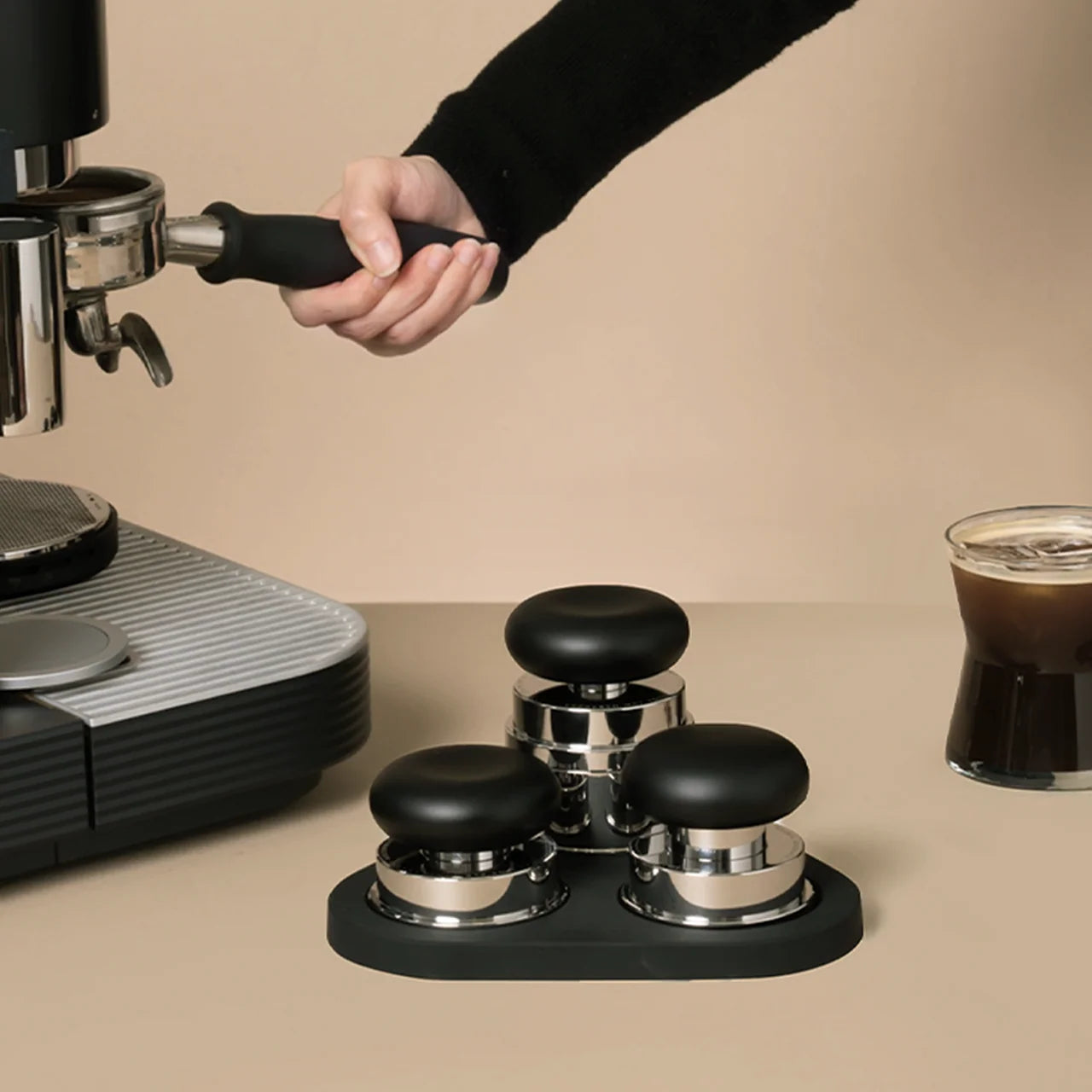 Person using an espresso machine with coffee grounds and a glass of coffee on a beige background